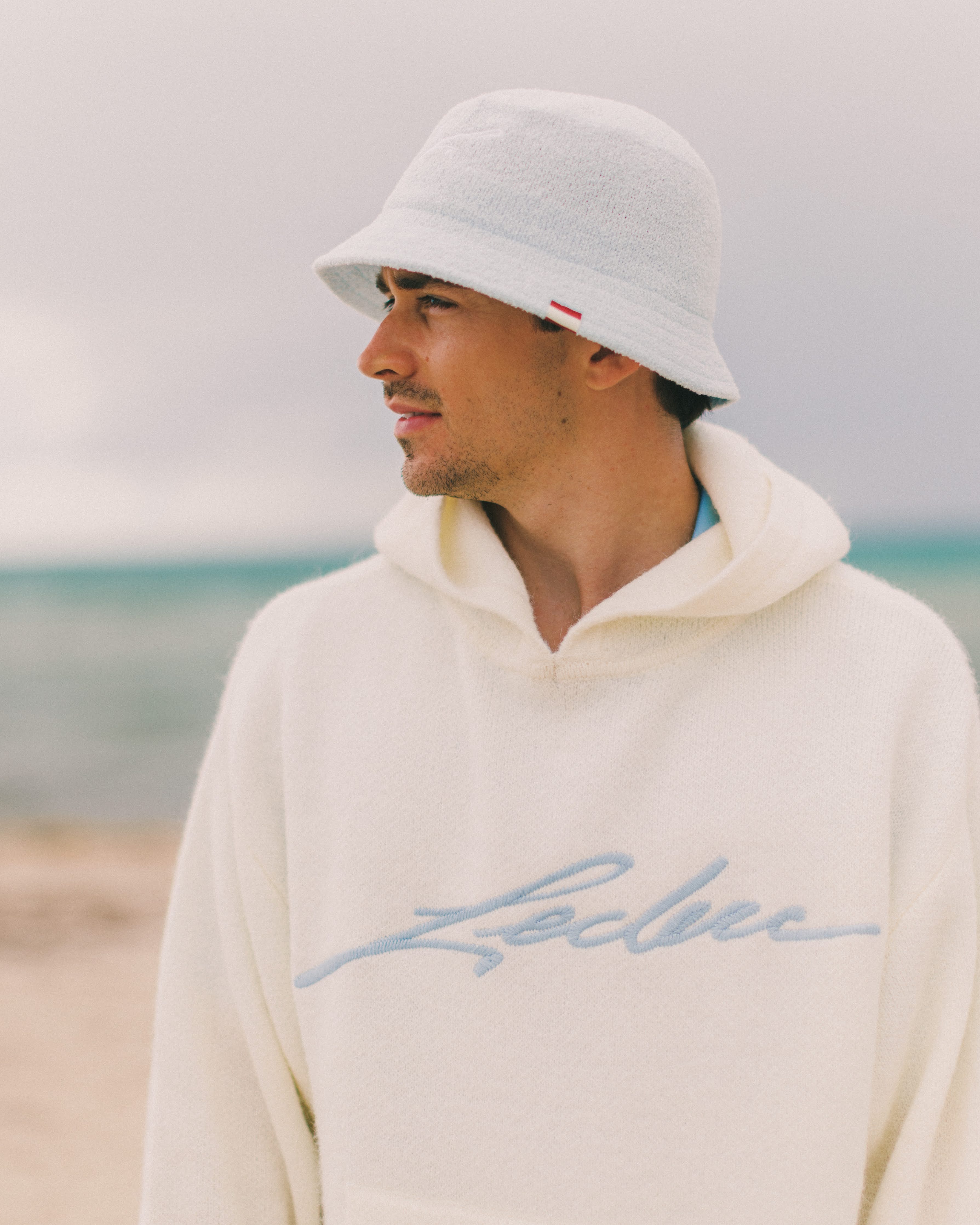 Charles Leclerc wearing a light-colored hoodie with 'Leclerc' branding on a beach.