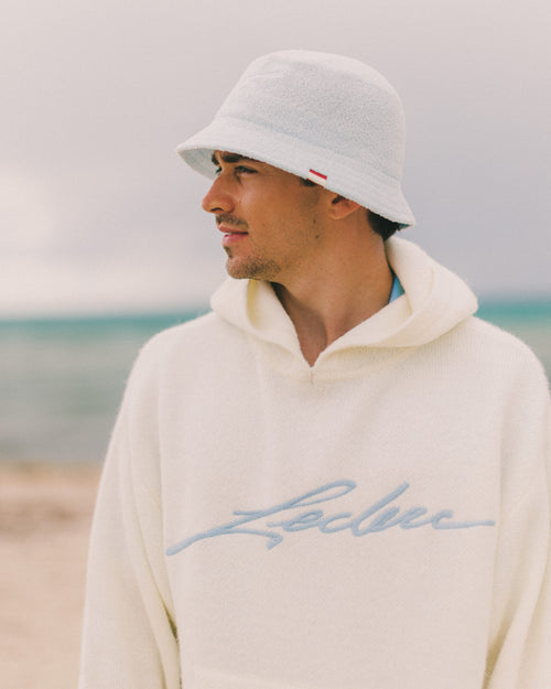 Charles Leclerc wearing a light-colored hoodie with 'Leclerc' branding on a beach.